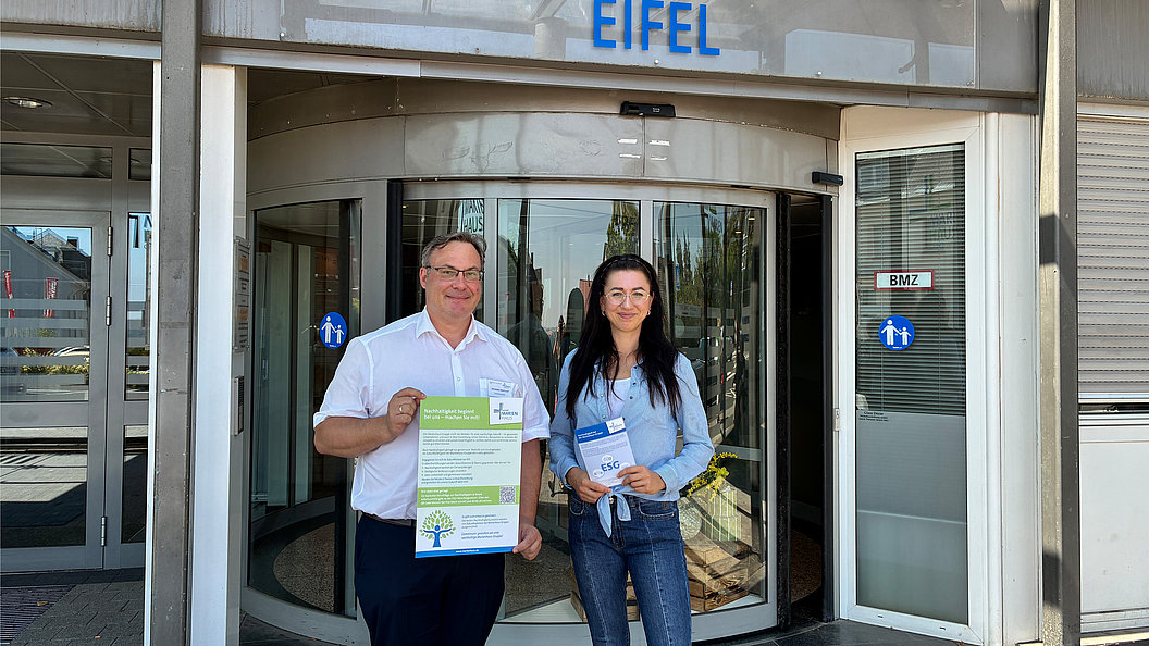Zwei Personen stehen vor einem Gebäude mit der Aufschrift 'EIFEL', der Mann hält ein Plakat, die Frau hält Broschüren.