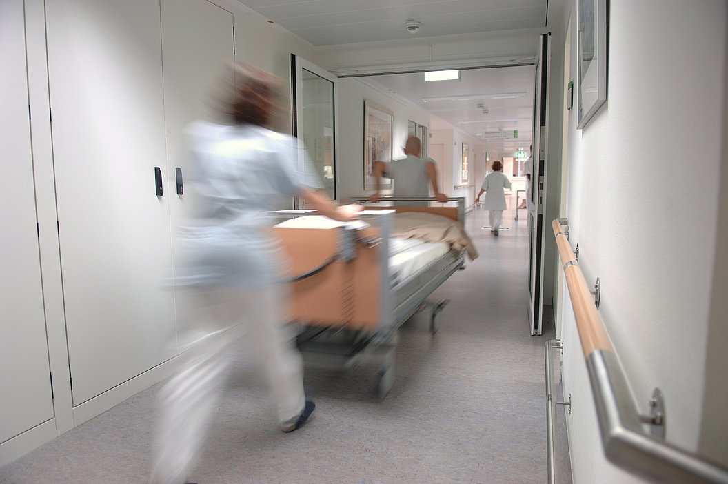 Krankenhausflur mit zwei Personen in weißer Kleidung, die ein Bett schieben, und einer weiteren Person am Ende des Flurs