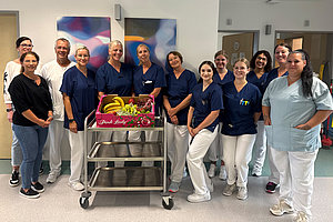 Gruppe von Menschen in medizinischer Kleidung steht in einem Flur um einen Wagen mit Obstkiste