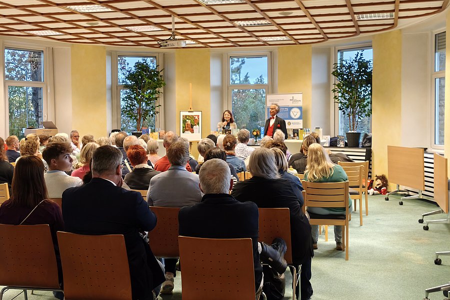 Publikum sitzt in einem hellen Raum mit großen Fenstern und Holzdecke und hört zwei Personen zu, die vorne sprechen