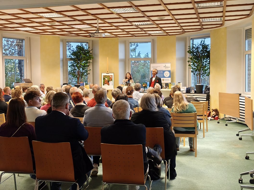 Publikum sitzt in einem hellen Raum mit großen Fenstern und Holzdecke und hört zwei Personen zu, die vorne sprechen