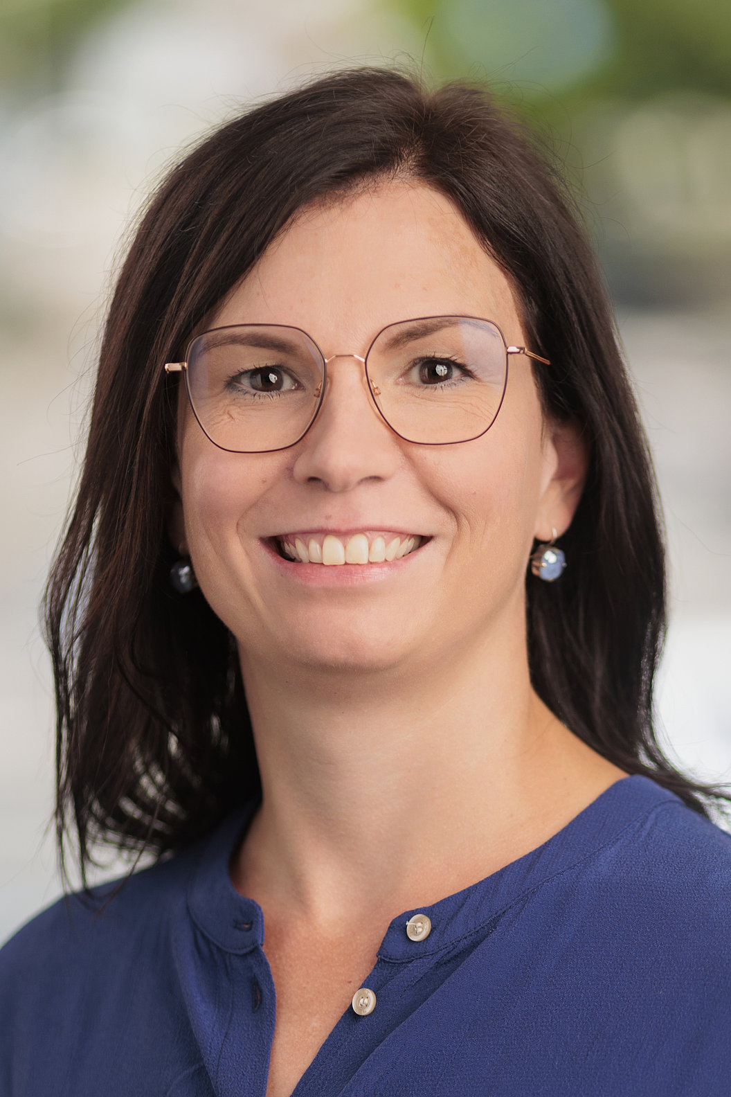 Frau mit schulterlangen braunen Haaren, roségoldenen Brillengestell und blauem Oberteil lächelt in die Kamera