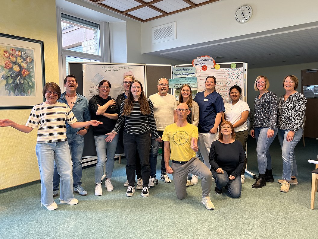 Gruppe von elf Personen in einem Raum mit Flipcharts und einer Uhr an der Wand, einige stehen, andere knien oder sitzen auf dem Boden