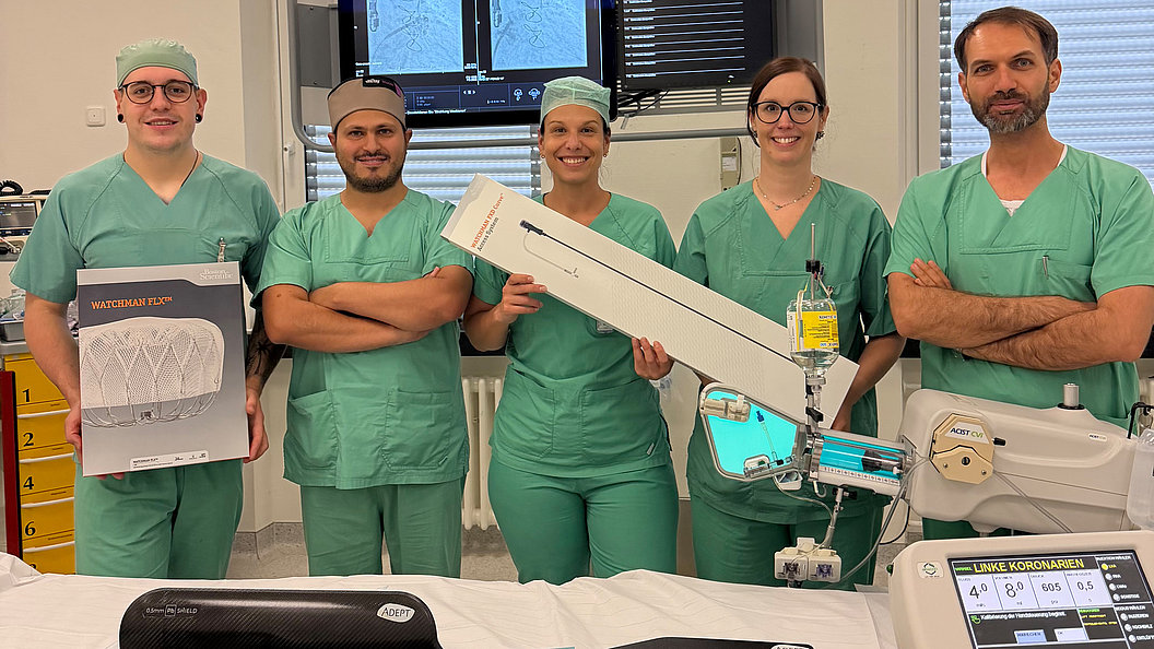 Fünf Personen in grüner OP-Kleidung stehen in einem Raum mit medizinischer Ausrüstung und halten zwei große Kartons mit medizinischen Geräten
