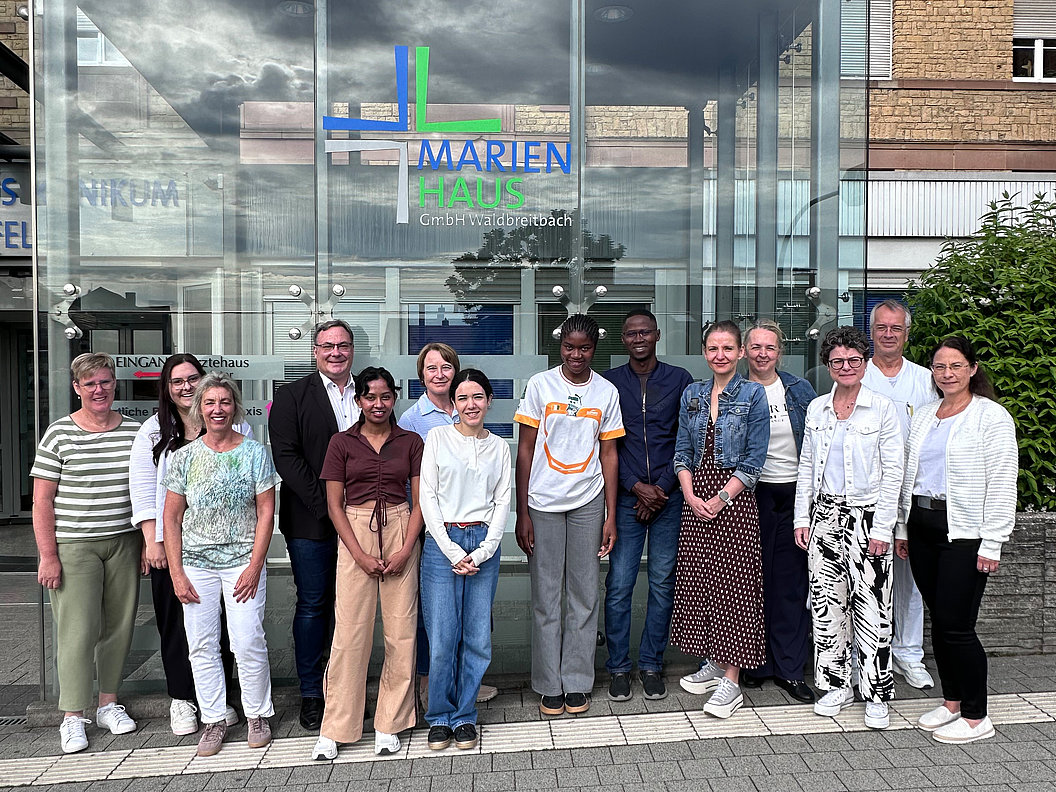 Gruppe von Menschen unterschiedlichen Alters und Geschlechts steht vor einem Gebäude mit Glasfassade und Schriftzug 'MARIEN HAUS GmbH Waldbreitbach'
