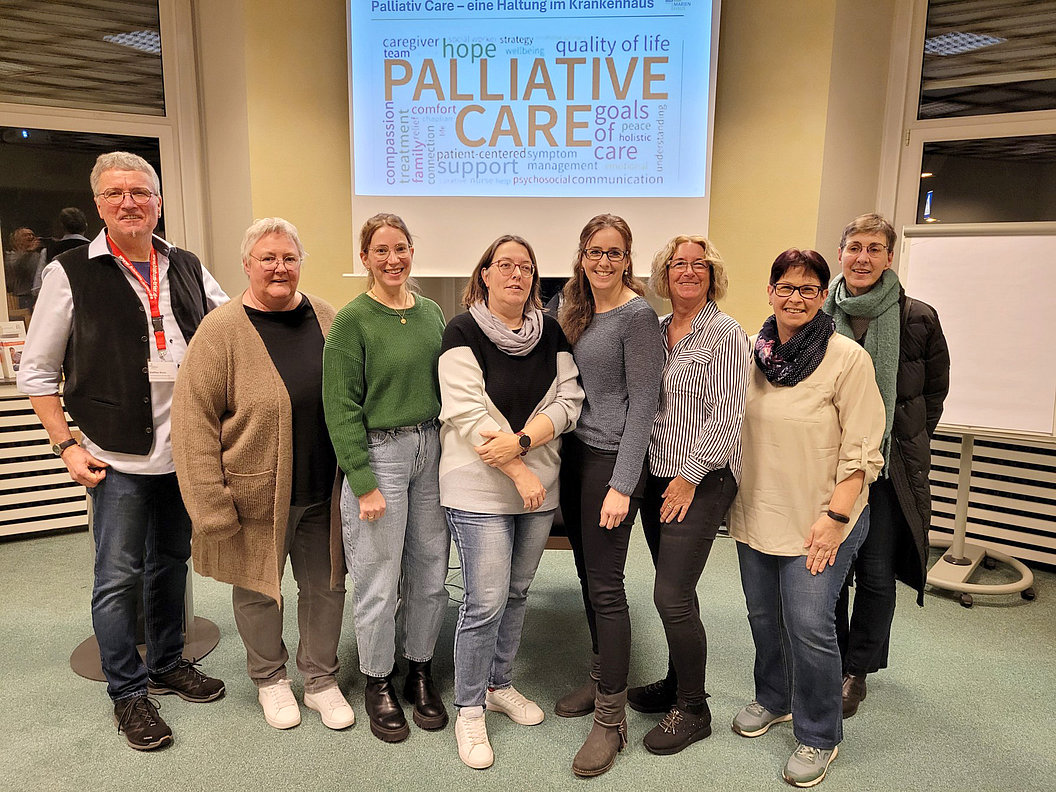 Gruppe von acht Personen steht vor einer Wand mit einer Projektion zum Thema Palliative Care in einem Raum mit Teppichboden und Rollstuhl im Hintergrund