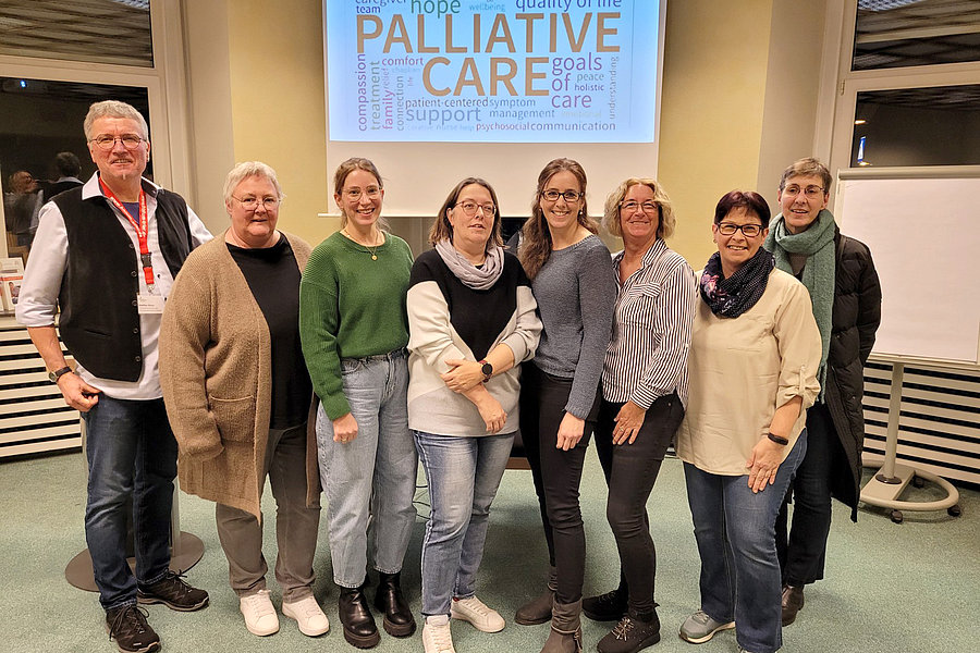 Gruppe von acht Personen steht vor einer Wand mit einer Projektion zum Thema Palliative Care in einem Raum mit Teppichboden und Rollstuhl im Hintergrund