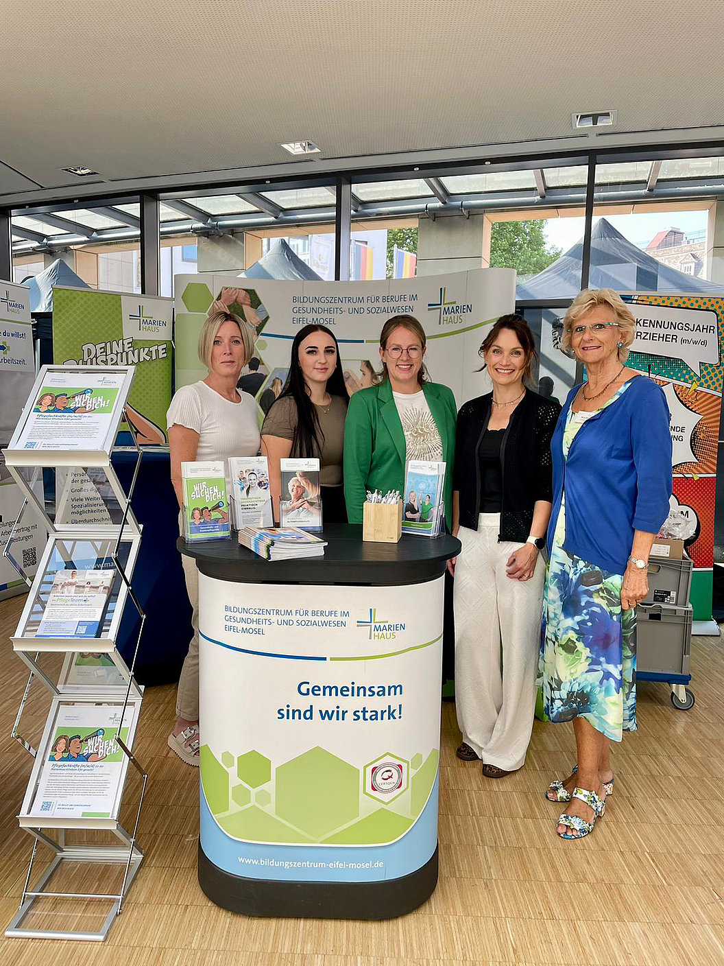 Fünf Frauen stehen an einem Messestand mit Informationsmaterial und Bannern zum Bildungszentrum für Berufe im Gesundheits- und Sozialwesen Eifel-Mosel
