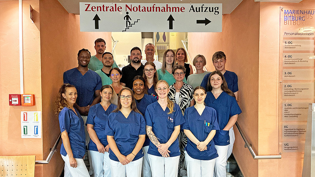 Gruppe von medizinischem Personal in blauen Kitteln steht in einem Krankenhausflur unter einem Schild mit der Aufschrift 'Zentrale Notaufnahme Aufzug'