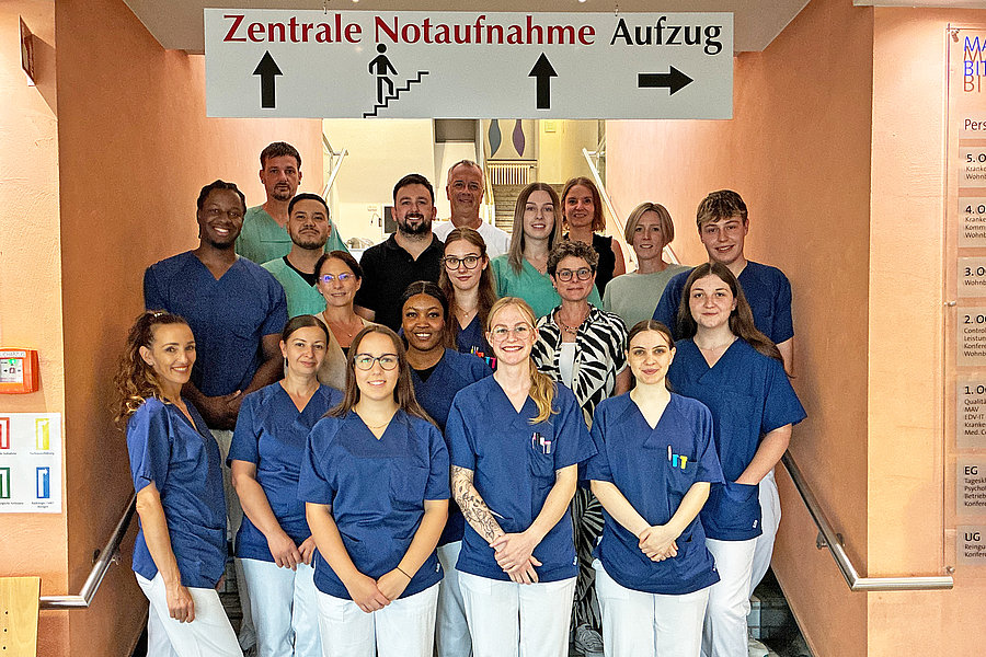 Gruppe von medizinischem Personal in blauen Kitteln steht in einem Krankenhausflur unter einem Schild mit der Aufschrift 'Zentrale Notaufnahme Aufzug'