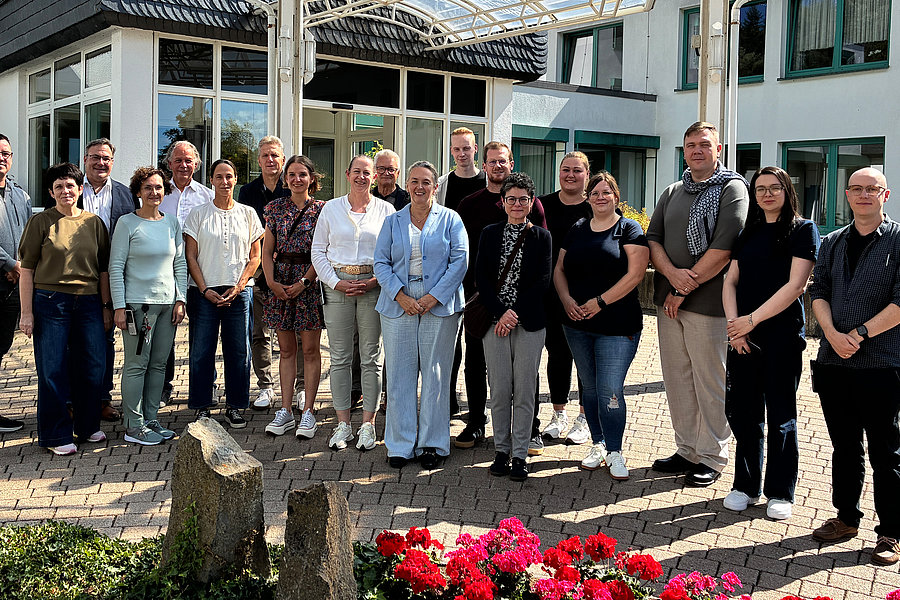 Gruppe von Menschen unterschiedlichen Alters steht vor einem Gebäude auf gepflastertem Hof bei Sonnenschein