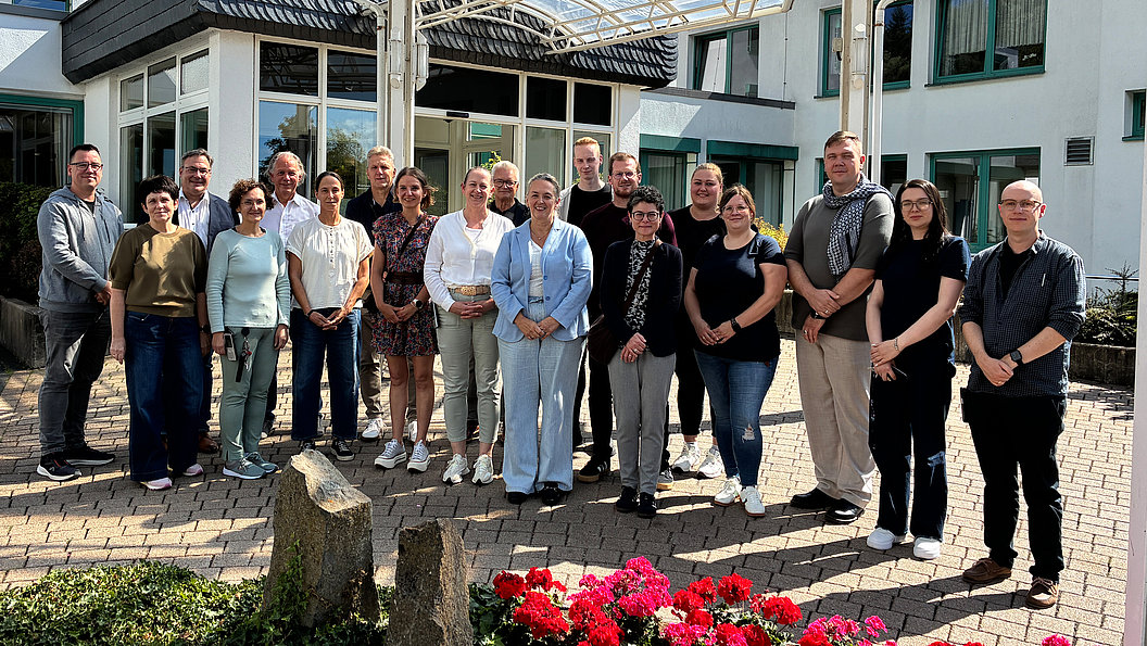 Gruppe von Menschen unterschiedlichen Alters steht vor einem Gebäude auf gepflastertem Hof bei Sonnenschein