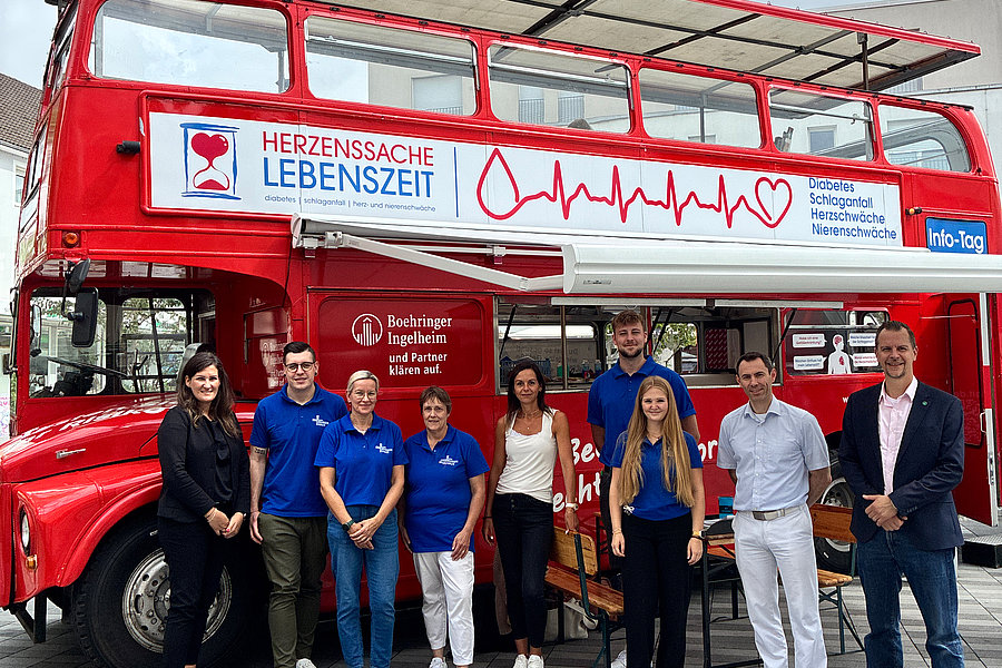 Rote Doppeldeckerbus mit Werbebanner "Herzenssache Lebenszeit" und Gruppe von acht Personen davor auf gepflastertem Platz