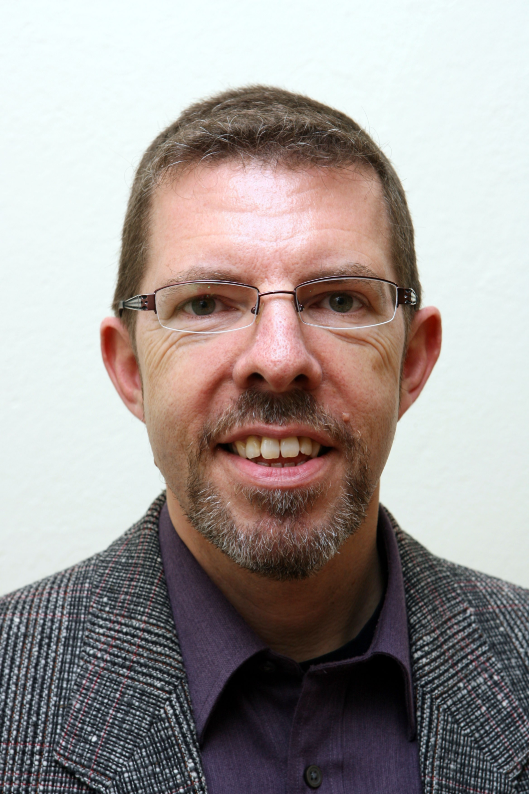 Mann mit Brille, kurzem braunem Haar und Bart trägt karierten Blazer und dunkles Hemd vor hellem Hintergrund